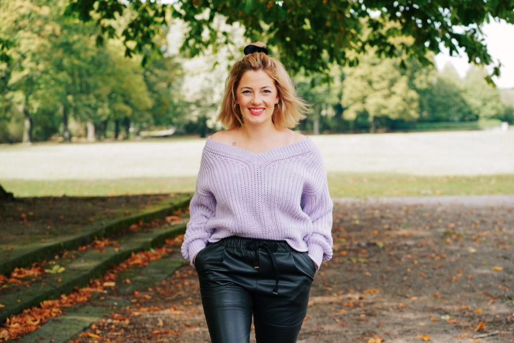 Angelina hat einen violetten Strickpullover und eine schwarze Hose an. Ihre blonden, schulterlangen Haare trägt sie offen. Sie geht lächelnd durch einen Park auf die Kamera zu.