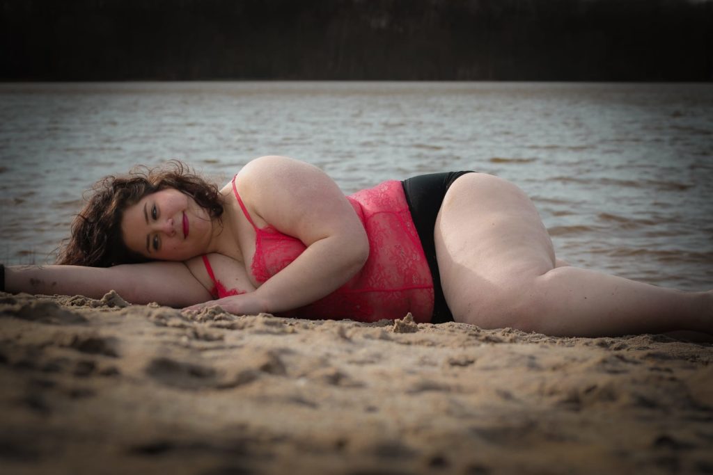 Livia liegt in einem roten Top und einer schwarzen Bikinihose an einem Strand und blick lächelnd in die Kamera.