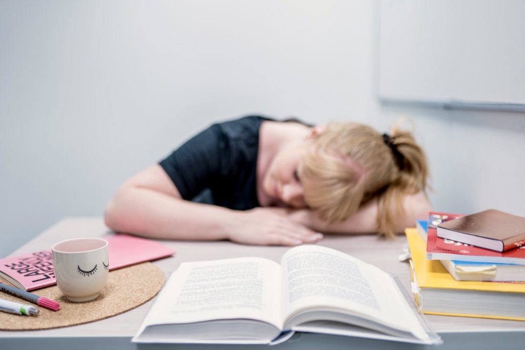 Ein blondes Mädchen hat den Kopf auf den Tisch gelegt. Vor ihr stapeln sich Bücher. Sie sieht erschöpft aus.