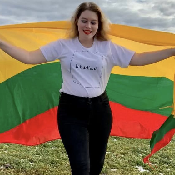 Die blonde Jessie steht auf einer Wiese. Hinter ihrem Rücken hält sie eine ausgebreitete Litauen-Flagge in den Farben Gelb, Grün und Rot