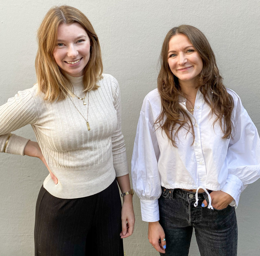 Elise und Lily tragen beide weiße Oberteile. Sie stehen vor einer weißen Wand und lächeln in die Kamera.