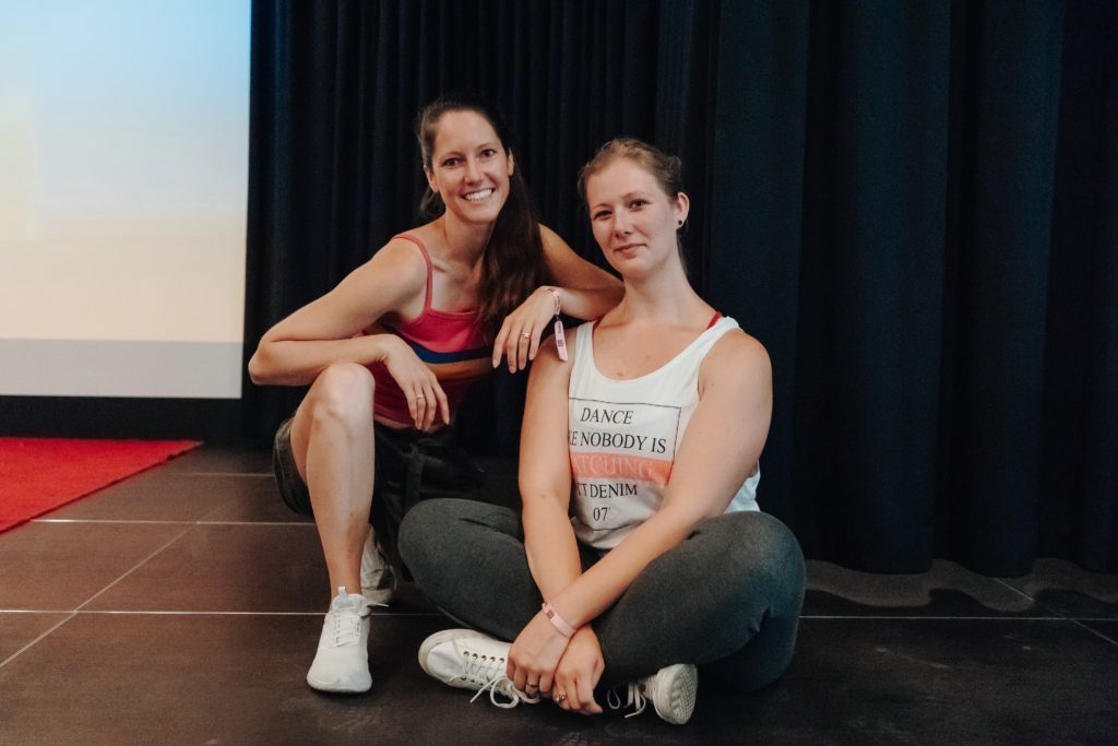 Sonja (links) und Linn (rechts), die Gründerinnen von ShedoesFuture sitzen auf einem dunklen Boden vor einem dunkelblauen Vorhang und lächeln in die Kamera