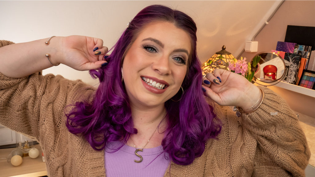 Sara hat ihre violetten Haare zu Locke frisiert. Sie lächelt in die Kamera und greift sich in die Harre.