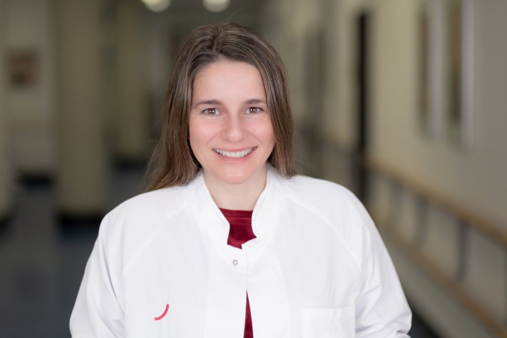 Dr. Mandy Mangler trägt einen weißen Ärztinnenkittel über einem roten Pullover. Sie steht auf einem Krankenhausflur und lächelt in die Kamera.