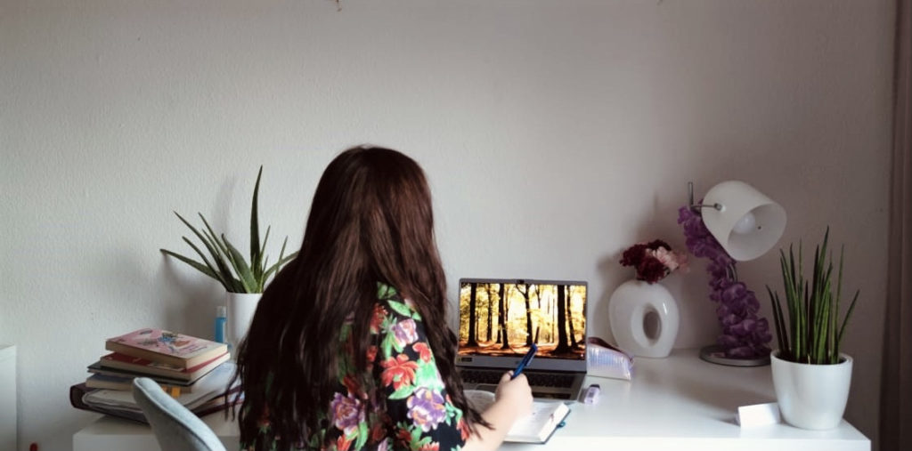 Ein brünettes Mädchen/ junge Frau sitzt mit dem Rücken zur Kamera vor einem Laptop. Neben ihr stapeln sich Bücher und einige Pflanzen schicken den Tisch.