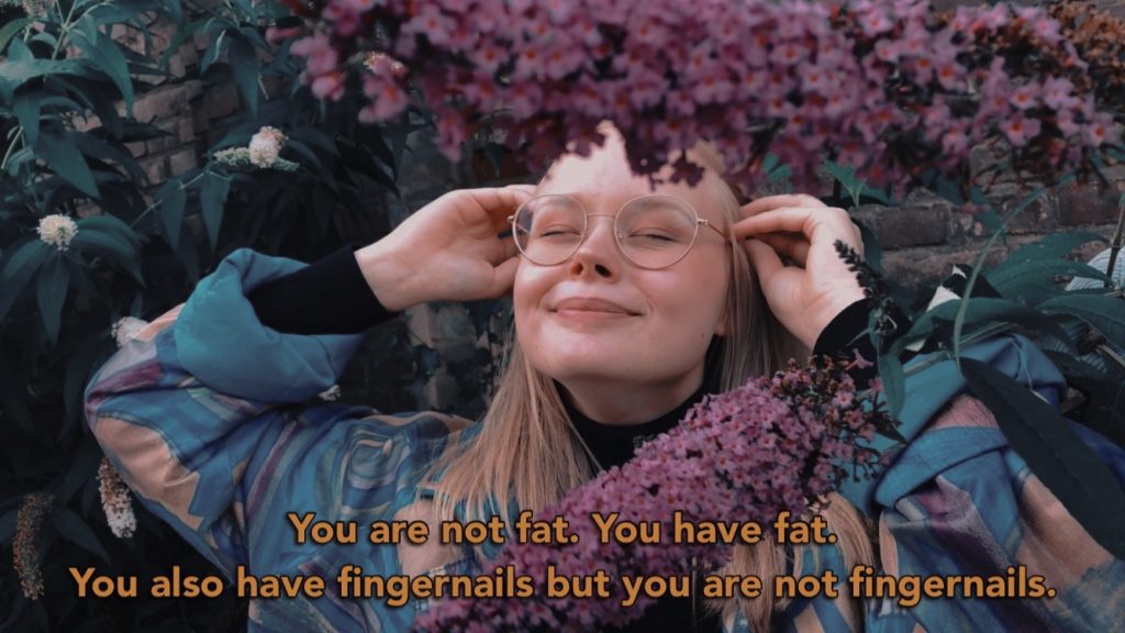 Marie steht vor einem Fliederbusch. Sie hat die Augen geschlossen und lächelt. Auf dem Foto steht "You are not fat. You have fat. You also have fingernails but you are not fingernails.