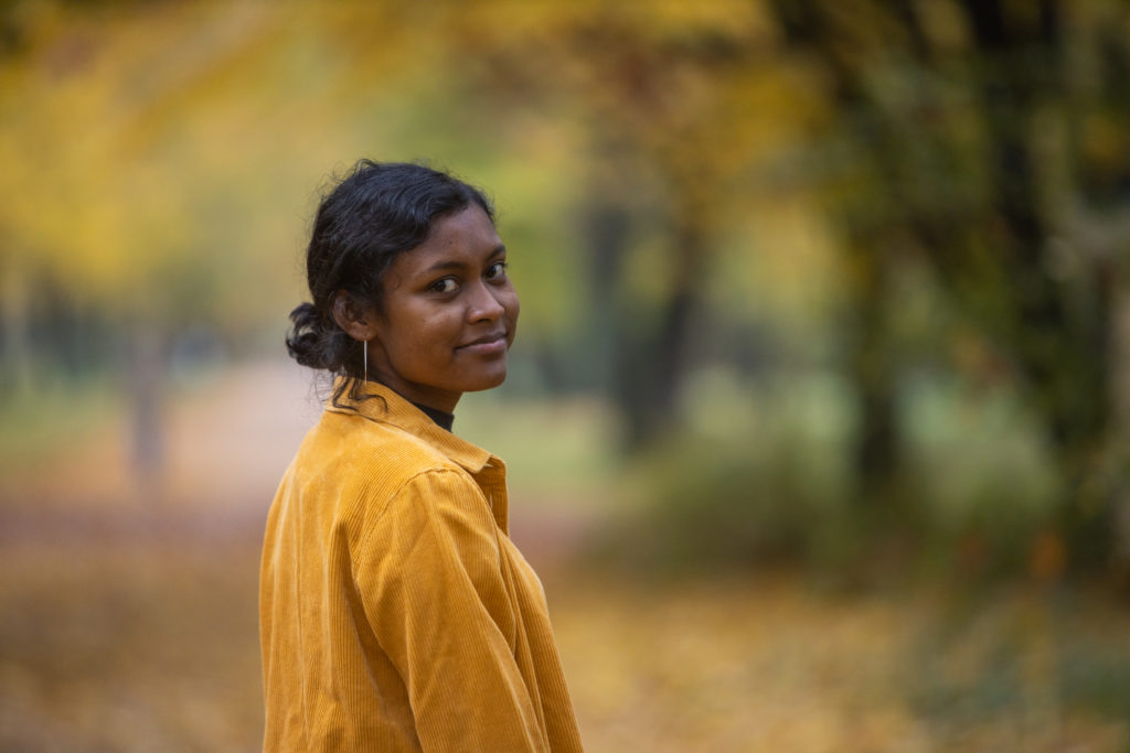 Sandhia steht in einem Herbswald. Sie trägt eine gelbe Jacke und blickt über ihre Schulter lächelnd in die Kamera