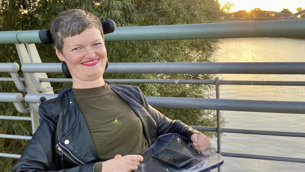 Anastasia sitzt in ihrem Rollstuhl auf einer Brücke. Hinter ihr geht über dem Wasser die Sonne unter