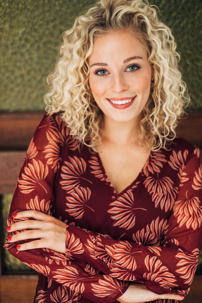 Charline wears a top with a red pattern and is standing in front of a green wall. Her arms are crossed in front of her body. She smiles in the camera.