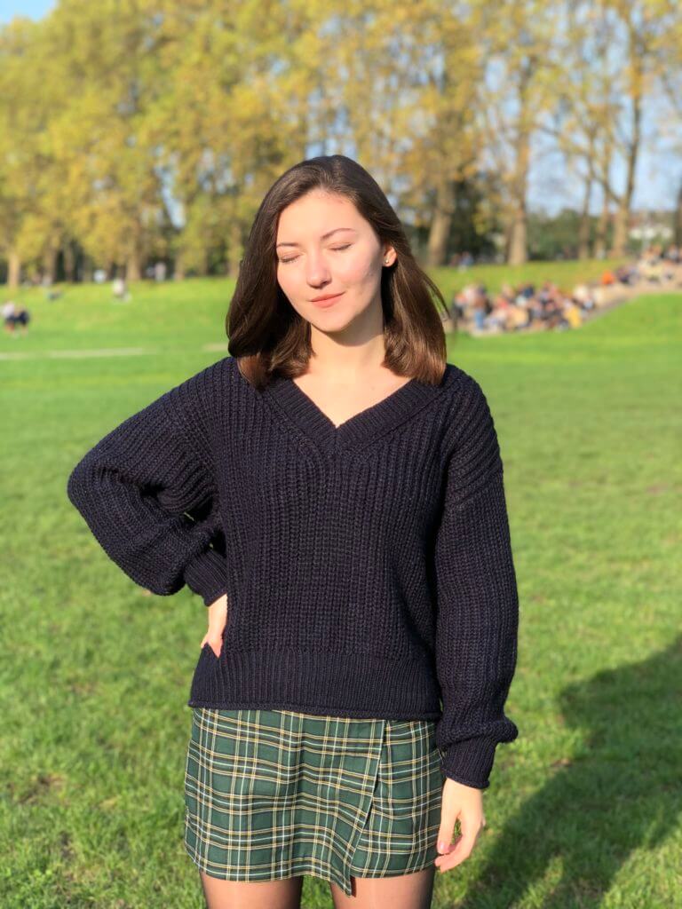 Nataliya trägt einen grün karierten Minirock und einen schwarzen Wollpullover. Sie steht in der Sonne in einem Park. Sie hat die Augen geschlossen.
