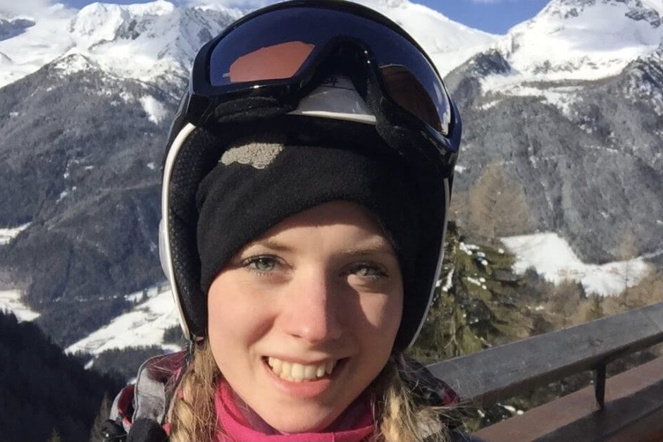 Ilona trägt einen Skihelm, auf den eine Skibrille gesetzt ist. Sie steht vor einem Bergpanorama und lächelt in die Kamera