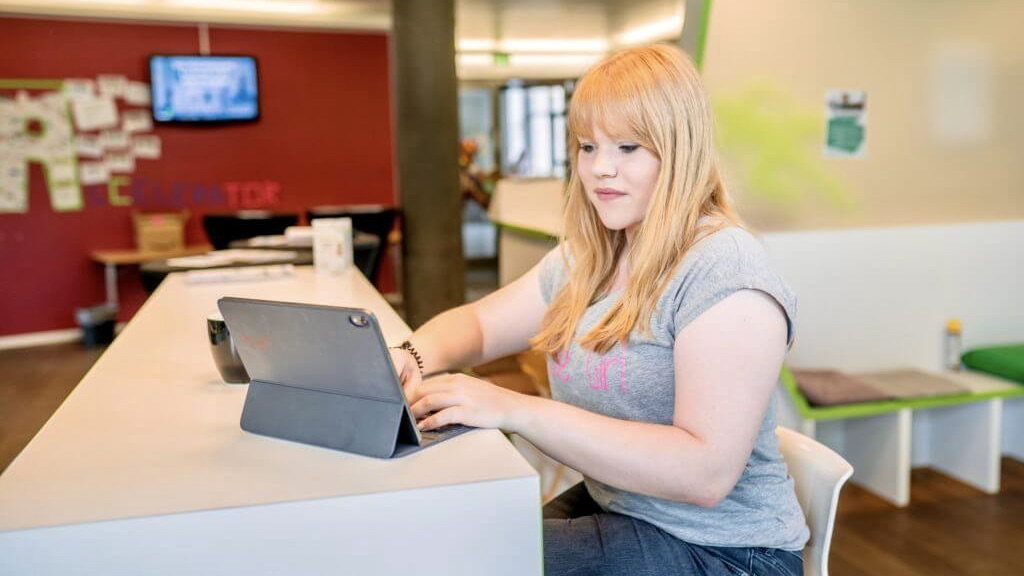 A young blonde woman is sitting in front of her laptop and is tiping.