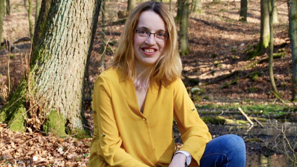 Karin sitz in einem herbstlichen Wald. Sie trägt eine blaue Jeans und eine gelbe Bluse und lächelt in die Kamera