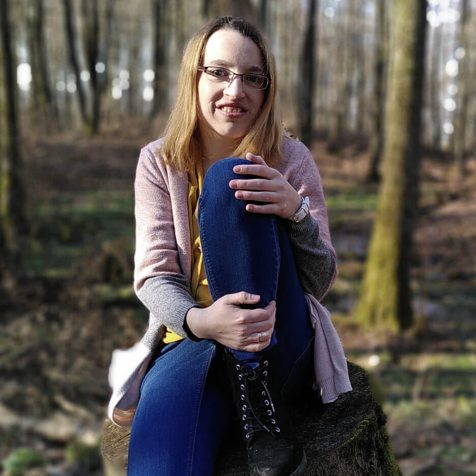 Karin sitzt auf einem abgesägten Baumstamm. Sie trägt eine blaue Hose und eine rosa Jacke und blickt in die Kamera
