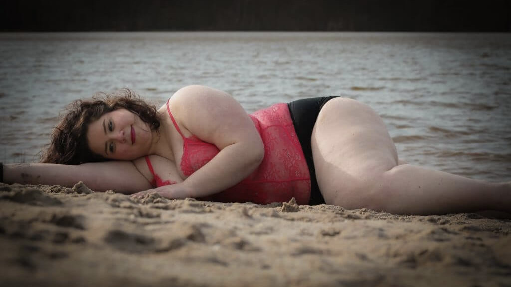 Livia liegt in einem roten Top und einer schwarzen Bikinihose an einem Strand und blick lächelnd in die Kamera.