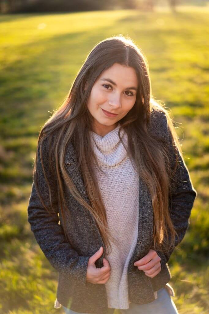 Maja trägt eine graue Jacke über einem hellen Wollpullover. Sie steht auf einer sonnigen Wiese und blickt in die Kamera
