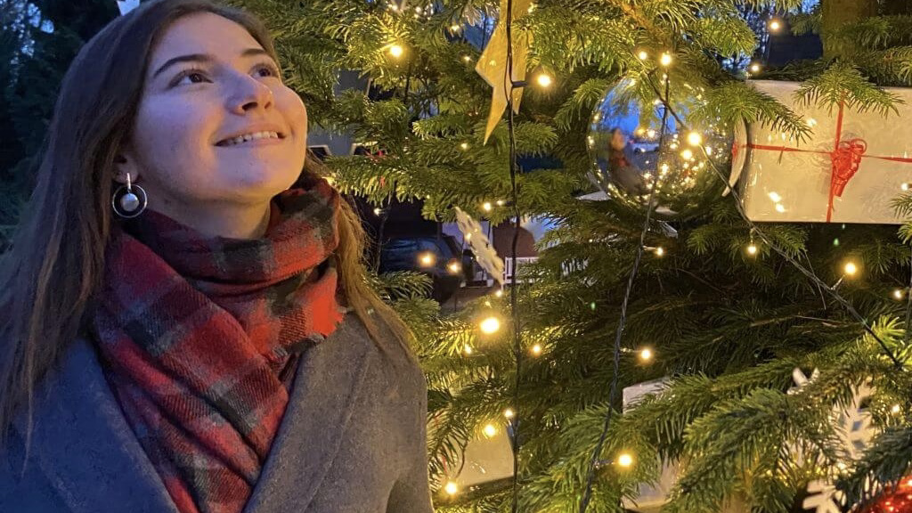 Nataliya steht vor einem weihnachtlich geschmückten und beleuchteten Tannenbaum. Sie blickt lächelnd nach oben
