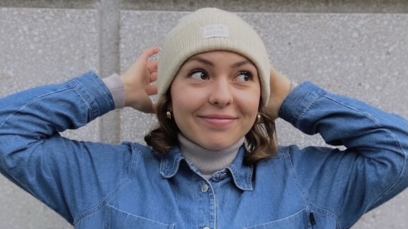 Nina wears a  denim shirt. Her brown hair is  shoulder-lenght and she is wearing a light coloured beanie.  Nina smiles into the camera.