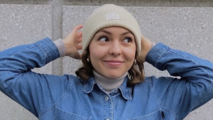 Nina trägt ein Jeanshemd. Ihre schulterlangen braunen Haare sind mit einer hellen Mütze bedeckt. Nina lächelt an der Kamera vorbei