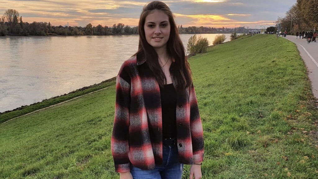 Tabitha trägt eine schwarz-rot karierte Jacke über einem schwarzen Top. Ihre dunklen Haare trägt sie offen. Sie steht auf einer Wiese mit dem Rücken zum Fluss und blickt in die Kamera