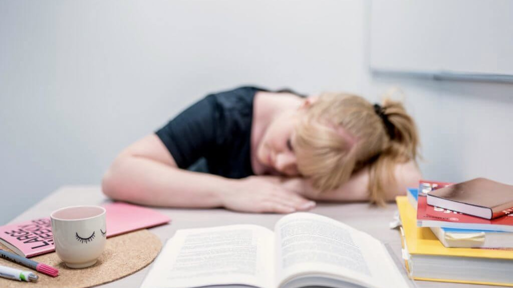 A blond girl is laying with her head on the table. The books are piling up in front of her. She looks exhausted. 