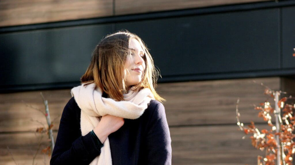 Eine brünette junge Frau trägt einen schwarzen Mantel und einen weißen Schal. Sie hat den Kopf Richtung Sonne gedreht.
